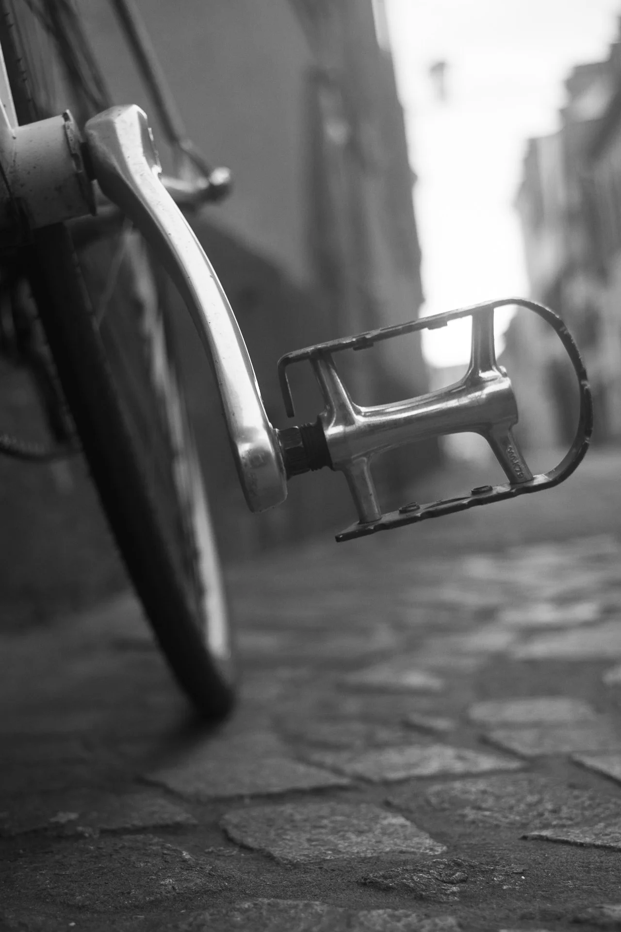 Black-and-white image of a bicycle pedal and crank arm, showing worn metal surface against cobblestone background. | Leica SL | N/A | f2.8 | 1/160s | ISO 100